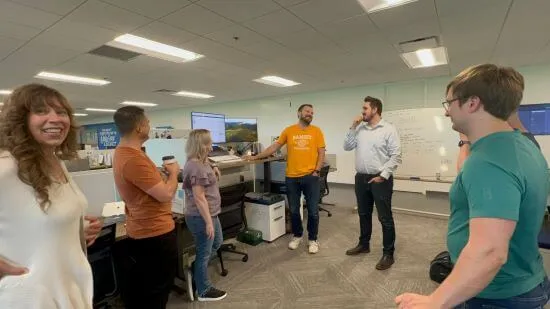 A photograph of the product squad in a standup meeting. Several team members are standing in a circle in the middle of an office.