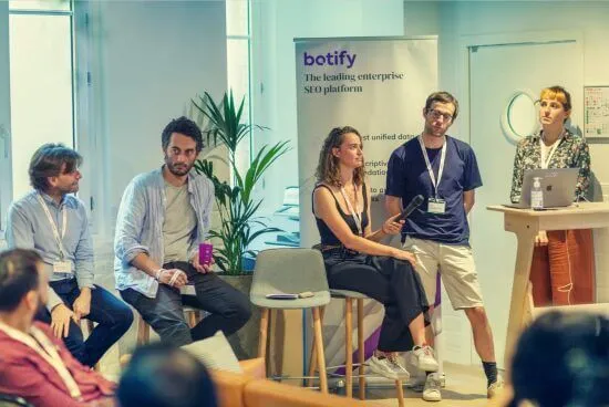 A photograph of Claire holding a microphone and sitting next to other Botify coworkers with other people gathered around.
