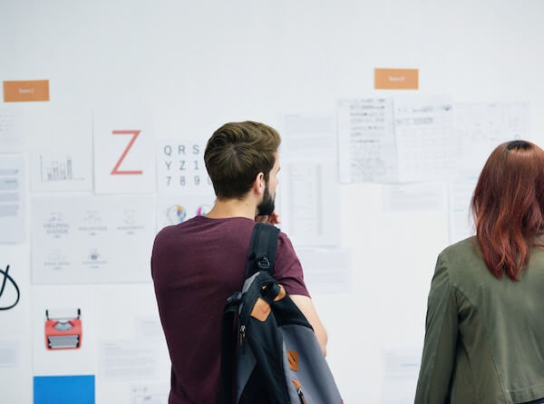 Two people standing looking at different text and images pasted on a whiteboard