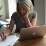 Rachel sits at a table with her laptop and printed out report, which she's reading and making notes on.