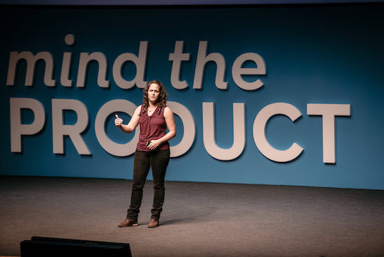 Teresa on stage in front of a large sign that says "Mind the Product." She has a serious expression on her face.