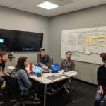 Lisa's team gathered around a table in a conference room. There's a whiteboard in the background covered in sticky notes.