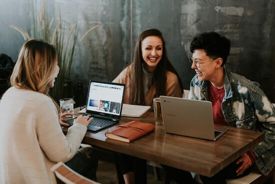 A group of people gathered around their laptops sitting together at a table and talking.