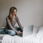 A woman is sitting on bed while working on a laptop.