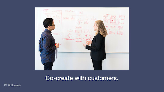 A photograph of two people standing in front of a whiteboard and talking to each other. The whiteboard appears to have some wireframes and user journey sketches on it. The caption reads, "Co-create with customers."