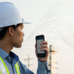 A man wearing a hard hat and safety vest is looking at some power lines while holding a mobile phone.