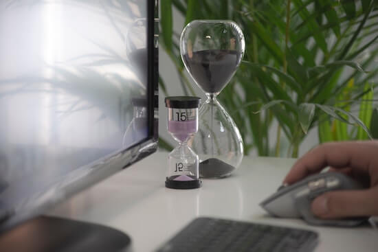 A photograph of a computer monitor and keyboard and a person's hand on a mouse. Next to the monitor there are two hourglass timers.