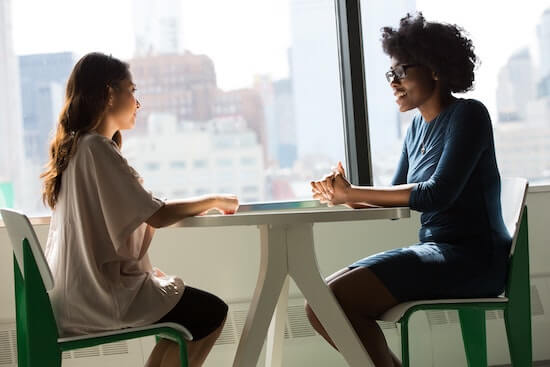 Two businesswomen are sitting at a table in an office talking to each other.