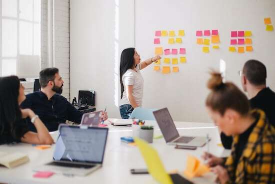 A photograph of a young woman standing in front of a whiteboard in a conference room. She is pointing to sticky notes on the whiteboard and speaking. The other people seated at the table are looking at her.