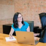 Two businesswomen sitting at a large table in an office. One of them is writing down notes on a notepad.