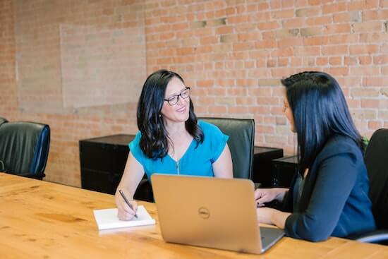 Two businesswomen sitting at a large table in an office. One of them is writing down notes on a notepad.