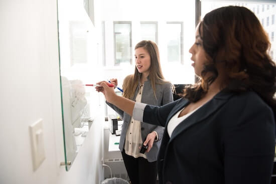 Two businesswomen standing in front of a whiteboard making notes on it.