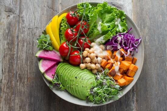 A photograph of a colorful bowl of salad full of avocado, sprouts, radishes, tomatoes, and sweet potatoes.