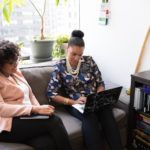 A photograph of two women sitting on a couch talking. One woman is taking notes on her laptop as they talk.