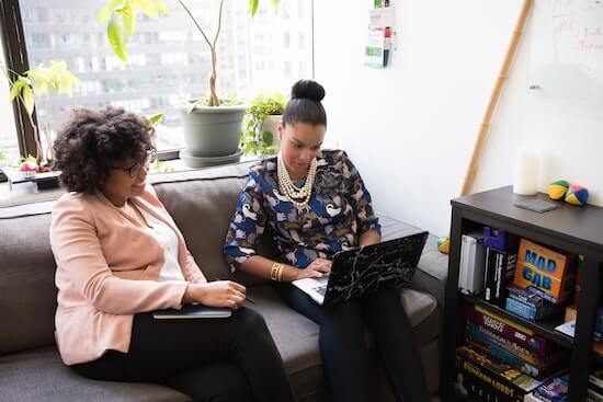 A photograph of two women sitting on a couch talking. One woman is taking notes on her laptop as they talk.