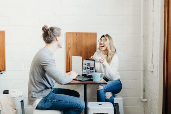 A photograph of two people sitting a table with laptops talking to each other and laughing.