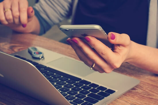 A photograph of a woman seated at a laptop holding a cell phone in her hand.