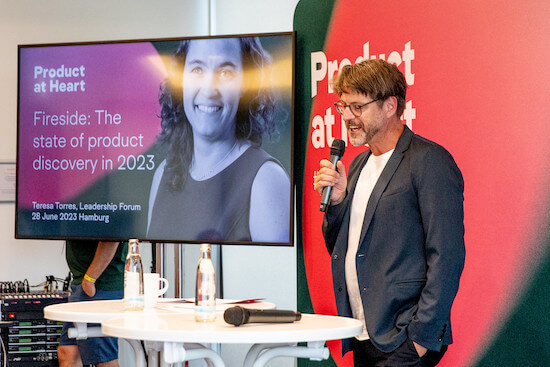 A photograph of Arne Kittler standing with a microphone in front of the "Fireside chat" slide at the Product at Heart conference.