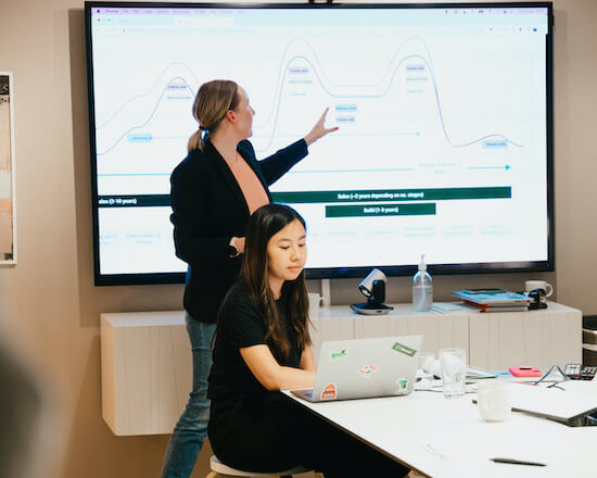 A photograph of two people in a conference room. One person is standing in front of a screen with a graph on it and the other is sitting at a table looking at a laptop.