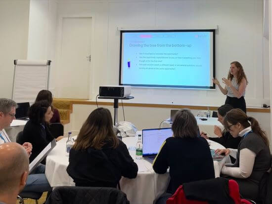 A photograph of Tali standing in front of a room of people while giving a presentation on opportunity solution trees.